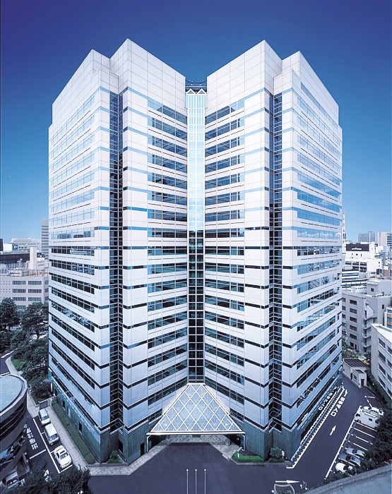 東京の中心地にある高層オフィスビル「虎ノ門ツインビルディング」。左右対称のデザインが特徴的で、中央にガラス張りのエントランスが配置されている。青空を背景に、白とガラスの外観が際立っている。